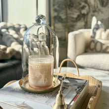 Load image into Gallery viewer, A clear glass cloche dome with a faceted top knob sits on a round gold tray, covering a textured cream candle. The setup rests on a stack of books and a woven tray in a softly lit living room.