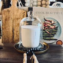 Load image into Gallery viewer, A white candle under a clear glass cloche dome, displayed on a brass tray with black beads and twine tassels, set on a dark wooden surface with a cookbook in the background.