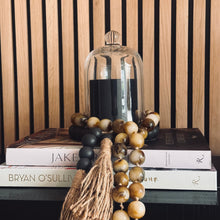 Load image into Gallery viewer, A decorative strand of large polished beads in earthy brown, tan, cream, and black tones with matching tassels, draped over a stack of books and a clear glass cloche against a wood slat wall.