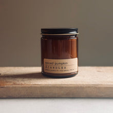 Load image into Gallery viewer, An amber glass jar candle with a black metal lid and a beige label reading 'spiced pumpkin' and 'ZCANDLES', placed on a rustic wooden board against a neutral taupe background.