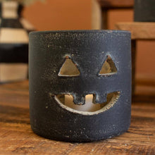 Load image into Gallery viewer, A black ceramic jack-o'-lantern candle holder with a textured finish, featuring cutouts for a face, sitting on a wooden table with a warm orange-brown background.
