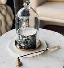 Load image into Gallery viewer, A glass cloche dome with a faceted knob lid covers a black candle with a white floral pattern and gold detailing, sitting on a white marble tray. A brass candle snuffer rests on the white marble table next to it, with a blurred living room background.
