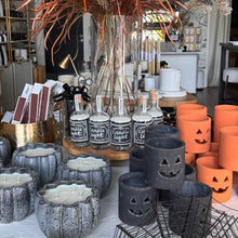Load image into Gallery viewer, A styled tabletop display of seasonal home decor and candles in a shop interior. The arrangement includes a large dried floral centerpiece in a tall glass vase, several clear bottles labeled 'Candle Light,' multiple pumpkin-shaped gray ceramic candle holders, orange cylindrical candle holders, and black jack-o'-lantern cutout candle holders.
