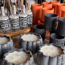 Load image into Gallery viewer, Pumpkin-shaped gray speckled ceramic candle holders filled with white wax candles, displayed alongside glass bottles labeled 'Candle Light' and orange candle sleeves.
