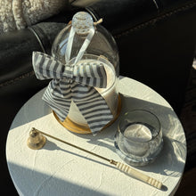 Load image into Gallery viewer, A decorative candle in a clear glass jar with a striped fabric bow and glass dome cover, styled on a white round table next to a small glass cup and a brass candle snuffer, with a dark leather sofa in the background.
