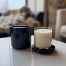 Load image into Gallery viewer, A black marble candle vessel with a white candle inside, placed on a white surface, with a blurred background featuring a cushion and part of a wall.
