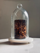 Load image into Gallery viewer, A clear glass cloche dome with a round knob top, displayed on a white marble base. Inside the dome is a cylindrical glass tumbler with a dark brown and amber mottled pattern resembling tortoiseshell.
