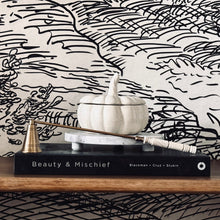 Load image into Gallery viewer, A styled tabletop scene featuring a black hardcover book titled 'Beauty &amp; Mischief' with a white pumpkin-shaped ceramic container and a brass candlestick on top, set against a black-and-white illustrated wallpaper background.
