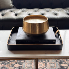 Load image into Gallery viewer, A large brass three-wick candle sits on a black book inside a black tray with gold trim, styled on a marble-top table in front of a tufted black leather sofa.
