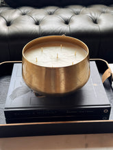 Load image into Gallery viewer, A large gold-toned metal bowl candle with multiple wicks sits on top of two stacked books in front of a black tufted leather sofa.
