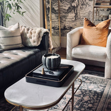 Load image into Gallery viewer, A styled living room corner featuring a black leather sofa, a cream armchair with a rust-orange pillow, a white marble coffee table with gold legs, and a black decorative tray holding a textured bowl. A tall floor lamp with a spherical white shade stands in the background against a black-and-white illustrated wallpaper and a bookshelf.
