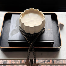 Load image into Gallery viewer, A cream-colored, scalloped ceramic candle holder containing a three-wick candle, styled on top of a black book and a dark wooden bead mala necklace, all resting on a black tray with a gold-trimmed edge.
