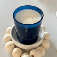 Load image into Gallery viewer, Blue candle in a glass holder on a white coaster with wooden beads.
