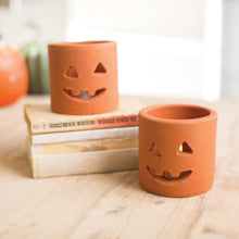 Load image into Gallery viewer, Two terracotta-colored ceramic candle holders shaped like jack-o'-lanterns, one standing on stacked books and one in front on a wooden surface, with a blurred orange pumpkin in the background.
